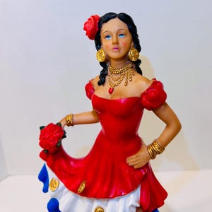Könnte beinhalten: Dekorative Figur einer Frau in einem roten Kleid, die einen Strauß roter Rosen hält. Die Figur hat schwarzes Haar, Goldschmuck und eine rote Blume im Haar. Das Kleid hat einen weißen und blauen Rock mit goldenen Akzenten.