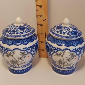 May include: Two matching blue and white ceramic jars with lids. The jars feature a floral and bamboo design with a bird. The lids have a white knob and a blue floral pattern. The jars are approximately 5 inches tall.