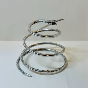 May include: A silver-toned metal object shaped like a coiled snake. The body of the snake forms a spiral structure with a textured surface that mimics scales. The head of the snake is positioned at the top of the coil, pointing outward.
