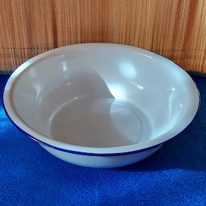 Könnte beinhalten: Eine weiße Emaille-Schüssel mit blauem Rand. Die Schüssel hat eine runde Form und eine glatte, glänzende Oberfläche. Die Schüssel ist leer und steht auf einer blau gemusterten Oberfläche.