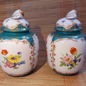 Puede incluir: Dos tarros de porcelana a juego con tapas. Los tarros son blancos con detalles en verde azulado y dorado. Diseños florales en rojo, amarillo, rosa y morado decoran el frente de cada tarro. Las tapas tienen un remate en forma de hoja.