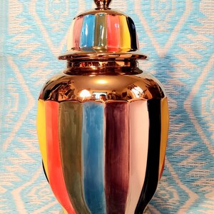 May include: A decorative ceramic jar with a lid. The jar is gold-colored with vertical stripes of various colors, including red, yellow, blue, and black. The lid has a gold finial and matching colorful stripes. The jar is approximately 12 inches tall.