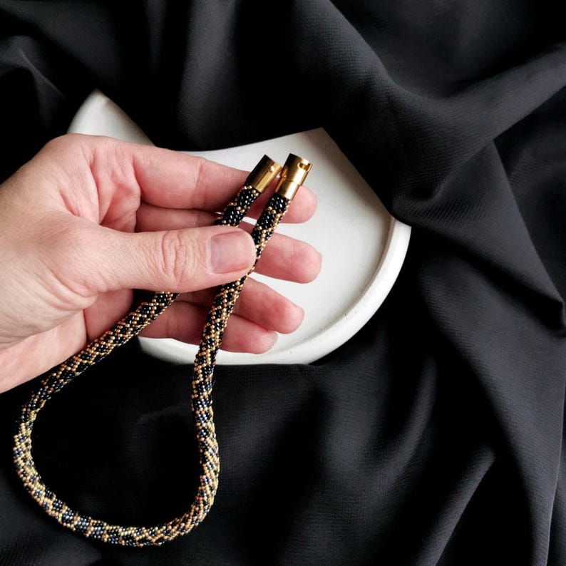 Op de afbeelding: Een zwarte en gouden kralenketting met goudkleurige sluitingen. De ketting is gemaakt van kleine, ronde kralen en heeft een geometrisch patroon.