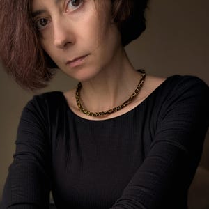 K&ouml;nnte beinhalten: Nahaufnahme einer Frau in einem schwarzen Langarmshirt und einer Perlenkette. Die Kette hat ein Muster aus schwarzen und goldenen Perlen. Die Frau hat kurze, rotbraune Haare und blickt direkt in die Kamera.