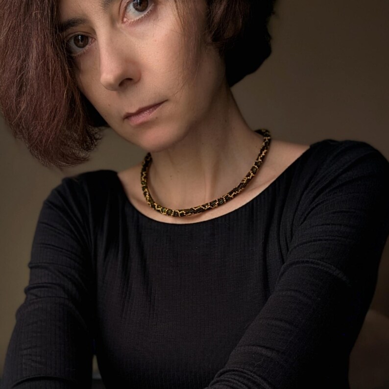 K&ouml;nnte beinhalten: Nahaufnahme einer Frau in einem schwarzen Langarmshirt und einer Perlenkette. Die Kette hat ein Muster aus schwarzen und goldenen Perlen. Die Frau hat kurze, rotbraune Haare und blickt direkt in die Kamera.