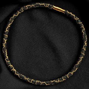 K&ouml;nnte beinhalten: Eine Halskette aus schwarzen und goldenen Perlen mit Zickzackmuster. Die Halskette besteht aus kleinen, runden Perlen und hat einen goldfarbenen Magnetverschluss. Pr&auml;sentiert auf schwarzem Hintergrund.
