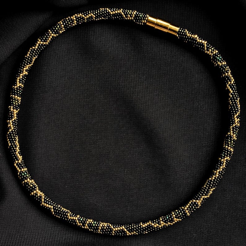K&ouml;nnte beinhalten: Eine Halskette aus schwarzen und goldenen Perlen mit Zickzackmuster. Die Halskette besteht aus kleinen, runden Perlen und hat einen goldfarbenen Magnetverschluss. Pr&auml;sentiert auf schwarzem Hintergrund.