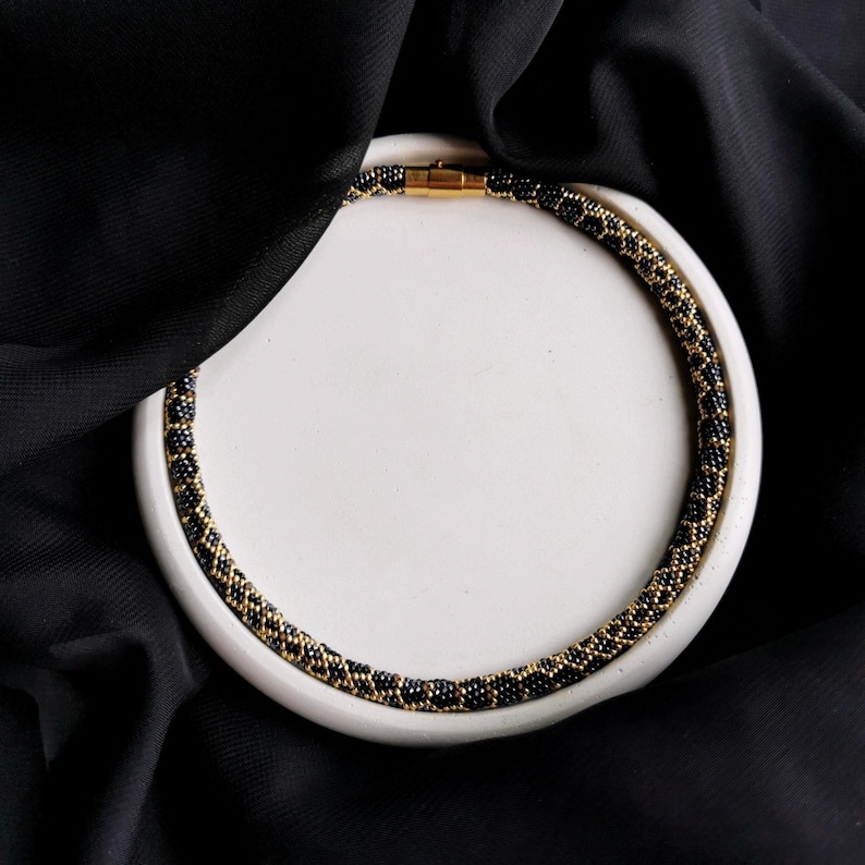 Op de afbeelding: Een zwarte en gouden kralenketting met een magneetsluiting, gepresenteerd op een wit rond oppervlak.