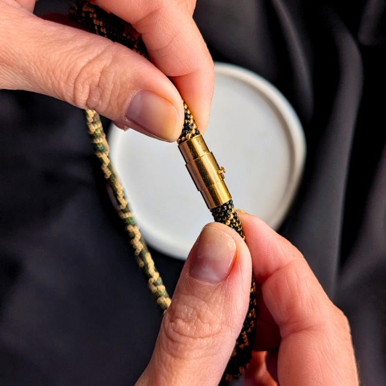 K&ouml;nnte beinhalten: Nahaufnahme einer Halskette mit einem schwarzen, gr&uuml;nen und goldenen Seildesign. Die Halskette hat einen goldfarbenen Verschluss. Der Verschluss wird von H&auml;nden gesichert. Der Hintergrund ist dunkel und einfarbig.