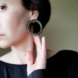 Peut inclure: Une paire de grandes boucles d'oreilles rondes en forme de cerceau, composées de perles noires et dorées. Les boucles d'oreilles sont portées par une personne aux cheveux foncés et portant une chemise noire.