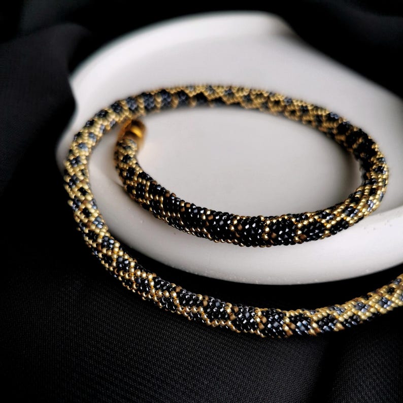 Op de afbeelding: Een zwarte en gouden kralenketting met een geometrisch patroon. De ketting ligt op een wit oppervlak.