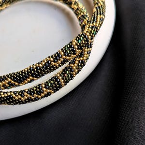 Peut inclure: Gros plan sur deux colliers de perles. Les colliers sont faits de petites perles noires et dor&eacute;es, cr&eacute;ant un aspect textur&eacute; en forme de corde. Les perles sont dispos&eacute;es en motif, avec des perles dor&eacute;es intercal&eacute;es parmi les perles noires. Les colliers reposent sur une surface blanche.