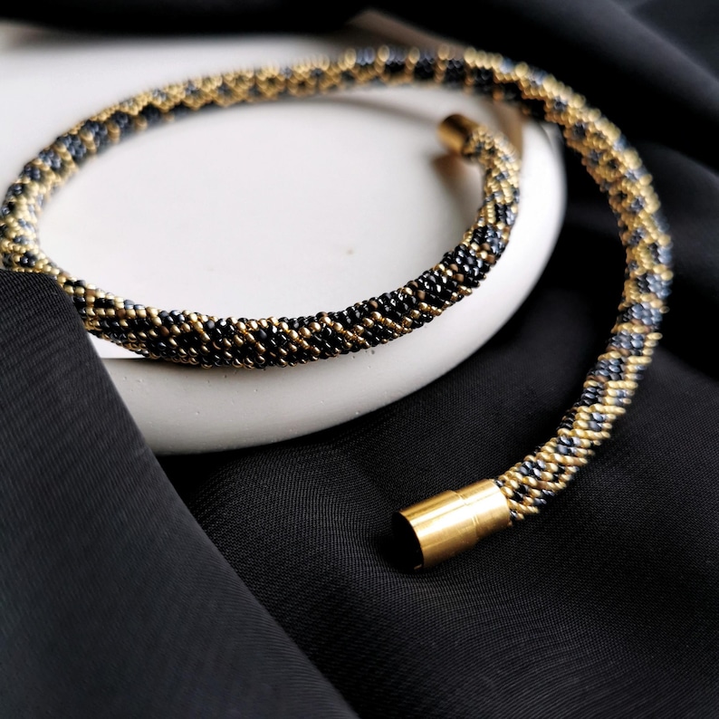 Op de afbeelding: Een zwarte en gouden kralenketting met een gouden sluiting. De ketting is gemaakt van kleine, ronde kralen die in een patroon zijn geweven.