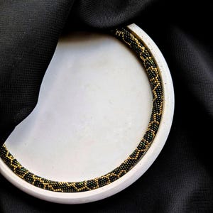 Peut inclure: Un collier de perles avec un motif noir et or repose sur une surface blanche et incurv&eacute;e. Le collier est compos&eacute; de petites perles rondes, cr&eacute;ant un aspect textur&eacute;. Le fond est un tissu sombre et textur&eacute;.