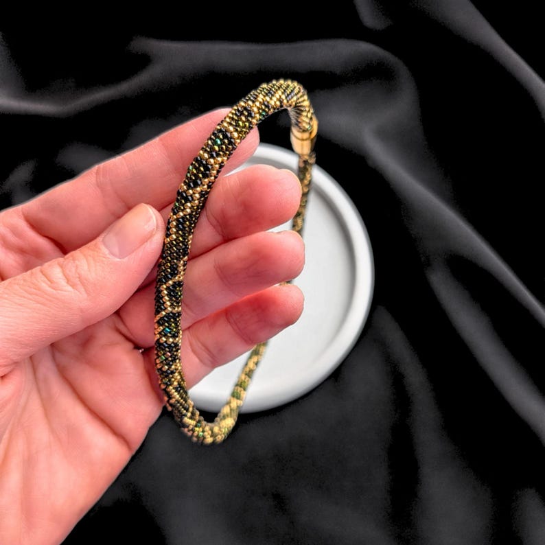 K&ouml;nnte beinhalten: Ein Perlenarmband mit einem goldenen Verschluss. Das Armband besteht aus kleinen, runden Perlen in Gold-, Schwarz- und Gr&uuml;nt&ouml;nen, die in einem sich wiederholenden Muster angeordnet sind. Das Armband wird vor einem schwarzen Hintergrund gezeigt.