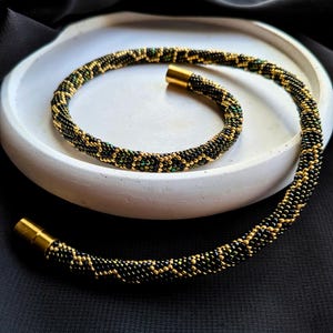 Pode incluir: Um colar de contas com um padrão preto, dourado e verde. O colar é exibido em uma bandeja branca e circular sobre um fundo preto. O colar tem fechos dourados.