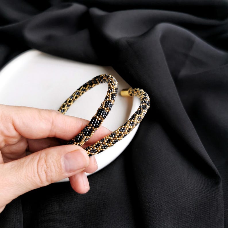 Op de afbeelding: Een paar grote creolen gemaakt van zwarte en gouden kralen. De oorbellen zijn rond en hebben een geweven patroon.