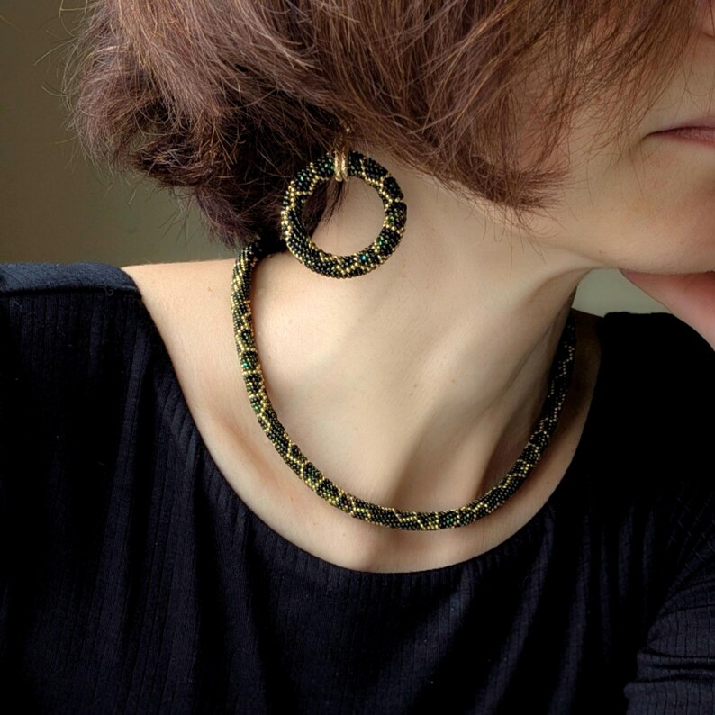 K&ouml;nnte beinhalten: Ein Schmuckset aus Perlen, bestehend aus einer Halskette und Creolen. Die Halskette und die Ohrringe sind schwarz, gr&uuml;n und goldfarben. Die Ohrringe sind kreisf&ouml;rmig, und die Halskette ist im Choker-Stil.