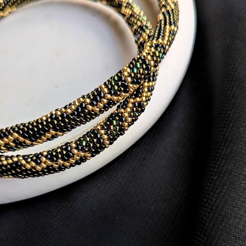 K&ouml;nnte beinhalten: Nahaufnahme von zwei Perlenketten. Die Halsketten bestehen aus kleinen schwarzen und goldenen Perlen, wodurch ein strukturiertes, seilartiges Aussehen entsteht. Die Perlen haben eine leicht irisierende Qualit&auml;t, die gr&uuml;ne und andere Farben reflektieren. Die Halsketten liegen auf einer wei&szlig;en Oberfl&auml;che vor einem schwarzen Hintergrund.