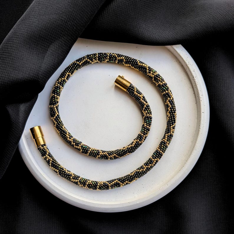 Peut inclure: Un collier de perles avec un motif noir, or et vert. Le collier est enroul&eacute; sur un plateau rond blanc sur un fond noir. Le collier a des fermoirs dor&eacute;s.