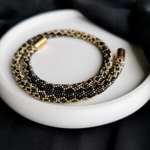 Op de afbeelding: Een zwarte en gouden kralenketting met een magneetsluiting. De ketting is gemaakt van kleine, ronde kralen die in een geometrisch patroon zijn gerangschikt.