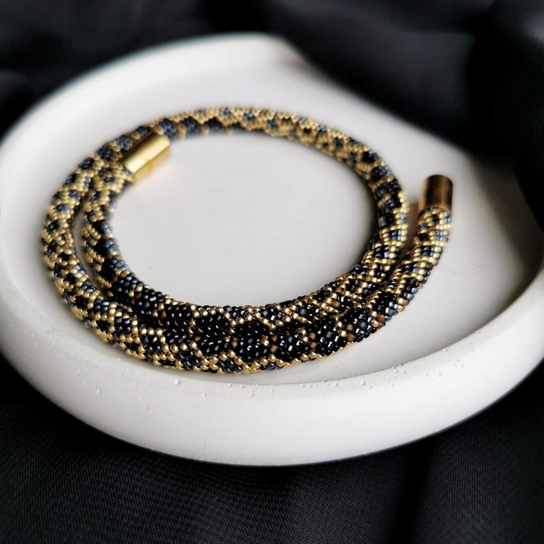 Op de afbeelding: Een zwarte en gouden kralenketting met een magneetsluiting. De ketting is gemaakt van kleine, ronde kralen die in een geometrisch patroon zijn gerangschikt.