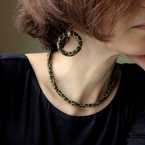 Peut inclure: Un ensemble de bijoux en perles comprenant un collier et des boucles d'oreilles cr&eacute;oles. Le collier et les boucles d'oreilles sont noirs, verts et dor&eacute;s. Les boucles d'oreilles sont circulaires et le collier est ras du cou.