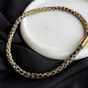 Op de afbeelding: Een zwarte en gouden kralenketting met een geometrisch patroon. De ketting is gemaakt van kleine kralen en heeft een magneetsluiting.