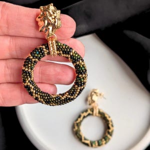 May include: Gold-toned earrings with a lion head design and beaded hoop. The hoops are black, gold, and green. The earrings are shown against a white surface and black background.