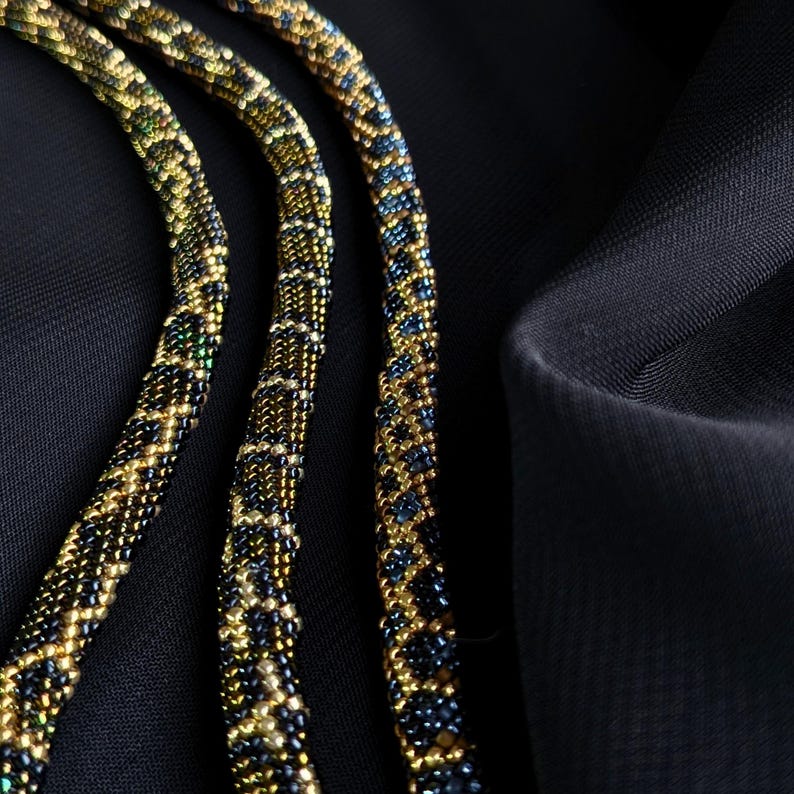 K&ouml;nnte beinhalten: Nahaufnahme von drei Perlenketten mit einem sich wiederholenden Muster aus goldenen, schwarzen und blauen Perlen. Die Halsketten sind auf einem schwarzen Stoffhintergrund angeordnet, was die aufwendige Perlenarbeit und das Design hervorhebt.