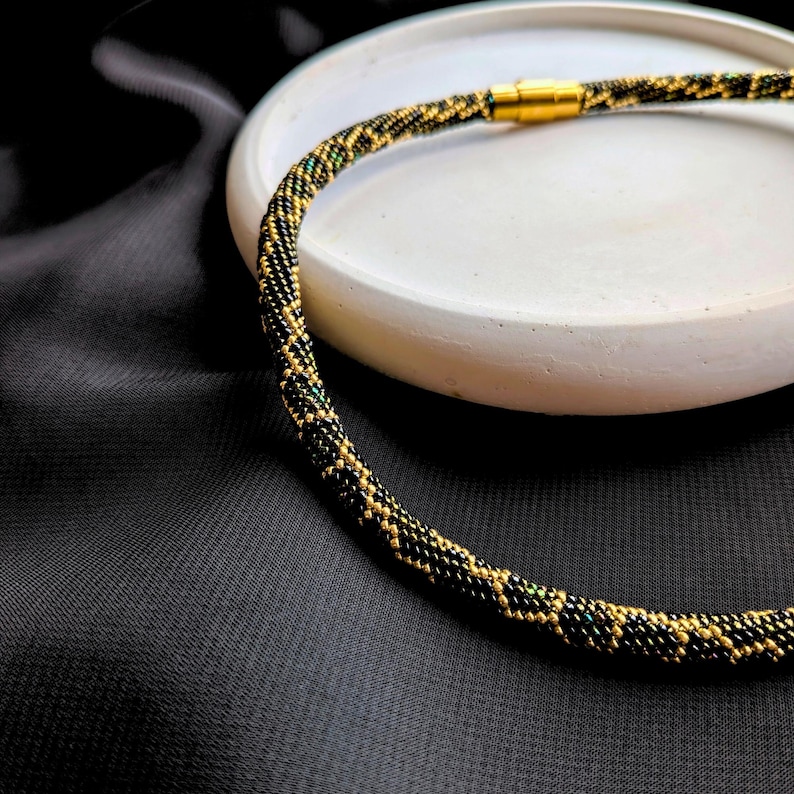 Peut inclure: Un collier de perles avec un fermoir magn&eacute;tique dor&eacute;. Le collier est principalement noir avec des accents dor&eacute;s et verts, cr&eacute;ant un motif textur&eacute;. Le collier est pr&eacute;sent&eacute; sur une surface ronde blanche sur fond noir.
