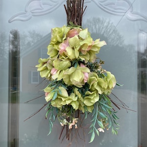 May include: A green and pink floral arrangement with a rustic twig base. The flowers are arranged in a cascading style, with the pink flowers at the top and the green flowers cascading down.