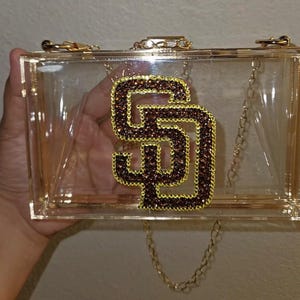 Pode incluir: Uma bolsa clutch retangular de acrílico transparente com ferragens de metal dourado e uma alça de corrente dourada removível. A frente apresenta um grande logotipo SD proeminente adornado com strass marrons e amarelos, criando um efeito brilhante.