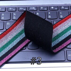 Pu&ograve; includere: Primo piano di un elastico nero con strisce argentate, rosa, verdi e rosse. L'elastico &egrave; piegato a met&agrave; e si trova su una tastiera di un laptop. Il testo #3 &egrave; visibile nell'immagine.