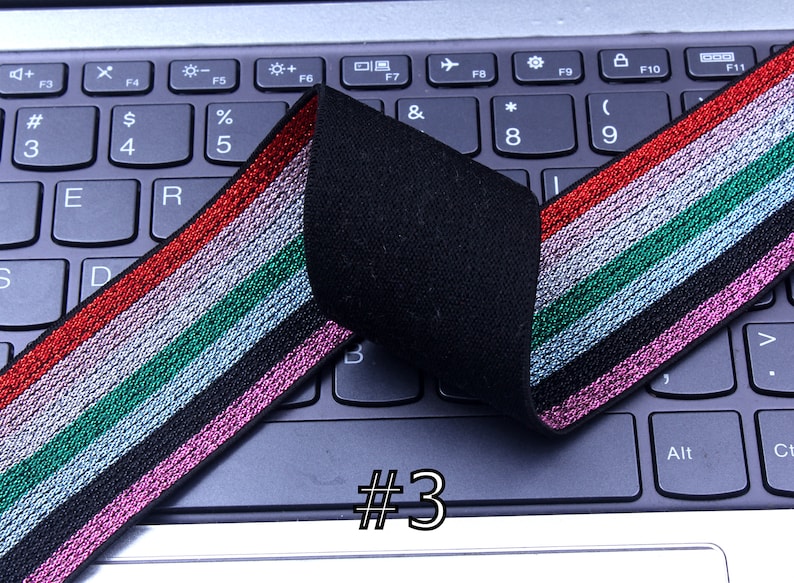 Pu&ograve; includere: Primo piano di un elastico nero con strisce argentate, rosa, verdi e rosse. L'elastico &egrave; piegato a met&agrave; e si trova su una tastiera di un laptop. Il testo #3 &egrave; visibile nell'immagine.