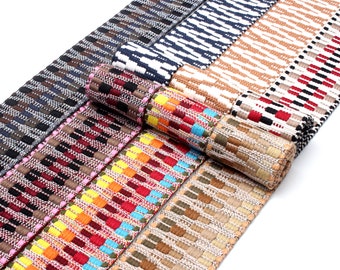 Könnte beinhalten: Sortiment an gewebten Stoffbändern in verschiedenen Farben und Mustern, darunter Braun, Schwarz, Weiß, Rot, Gelb, Orange, Blau und Grün. Die Bänder sind aufgerollt und in einer Reihe angeordnet.
