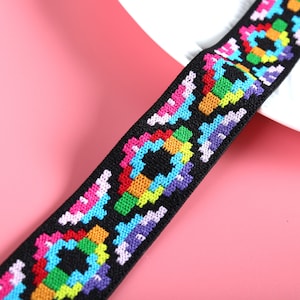 May include: A black elastic band featuring a repeating geometric pattern embroidered with vibrant thread colors including red, orange, yellow, green, blue, purple, and pink. The woven design consists of pixelated shapes arranged in a sequence.