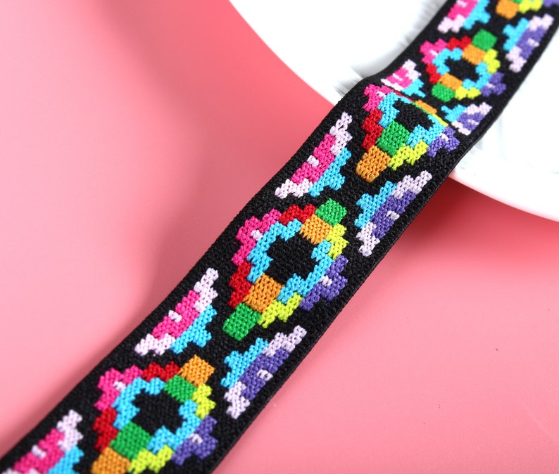 May include: A black elastic band featuring a repeating geometric pattern embroidered with vibrant thread colors including red, orange, yellow, green, blue, purple, and pink. The woven design consists of pixelated shapes arranged in a sequence.