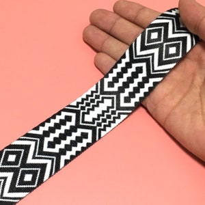 Könnte beinhalten: Schwarz-weiß gemusterter Gurt mit geometrischen Designs. Der Gurt hat ein sich wiederholendes Muster aus Rauten und Zickzacklinien. Der Gurt wird vor einem rosa Hintergrund gezeigt.