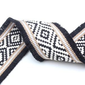 May include: Black and white geometric patterned trim with a fringe edge. The trim has a tan stripe running through the center.