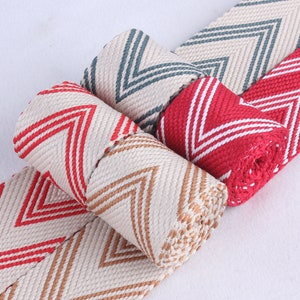 Może przedstawiać: Trzy rolki tkanej wstążki z wzorem chevron. Wstążka jest dostępna w kolorze czerwonym, beżowym i niebieskim.
