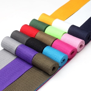 May include: A variety of colors of cotton webbing, rolled up and displayed on a white surface. The colors include gray, purple, green, blue, pink, red, black, white, and yellow.