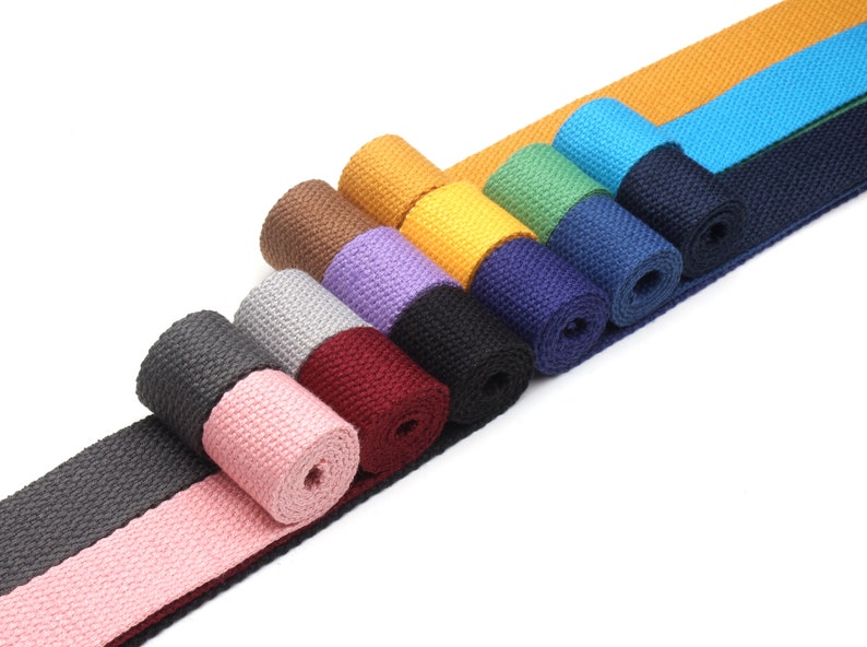 May include: A collection of 12 rolls of cotton webbing in various colors, including pink, gray, black, burgundy, purple, blue, green, yellow, and brown. The webbing is arranged in a diagonal line, with the ends of each roll overlapping.