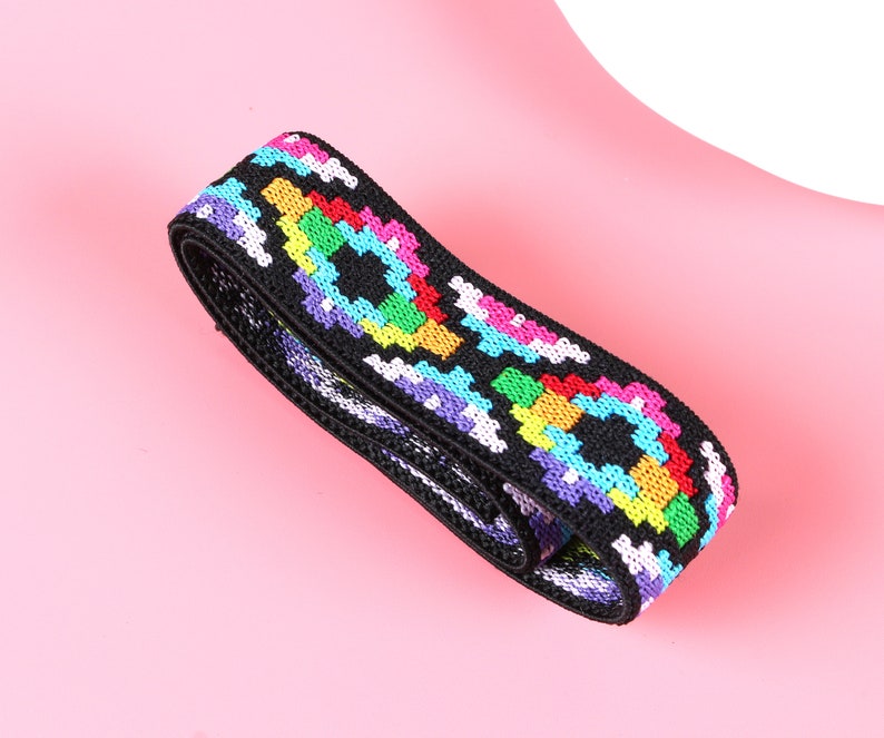 May include: A black elastic band featuring a repeating geometric pattern in vibrant colors including neon pink, bright yellow, turquoise, and purple. The woven fabric has a textured surface and is coiled into a loop, displaying a consistent design.
