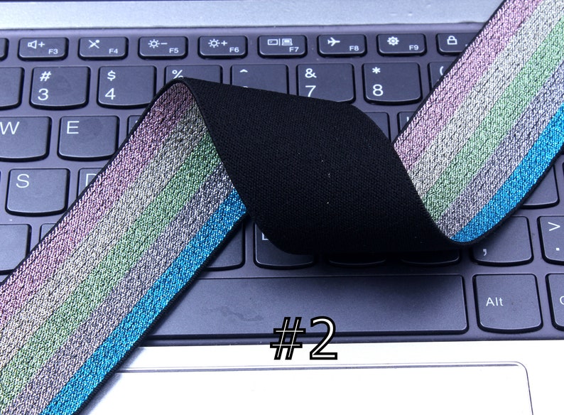 Pu&ograve; includere: Primo piano di un elastico nero con una striscia iridescente multicolore. L'elastico &egrave; piegato a met&agrave; e posizionato su una tastiera di un laptop. Il numero "#2" &egrave; scritto sulla tastiera.