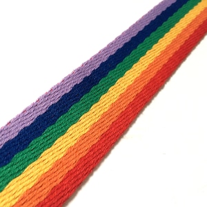 May include: A diagonal strip of rainbow-colored woven fabric. The colors, from top to bottom, are purple, blue, green, yellow, orange, and red. The fabric is against a white background.