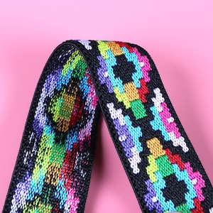 May include: A black elastic strap featuring a woven, multi-colored geometric pattern in vibrant shades of neon pink, bright blue, lime green, yellow, and purple. The fabric has a textured, woven appearance with a repeating, symmetrical design.