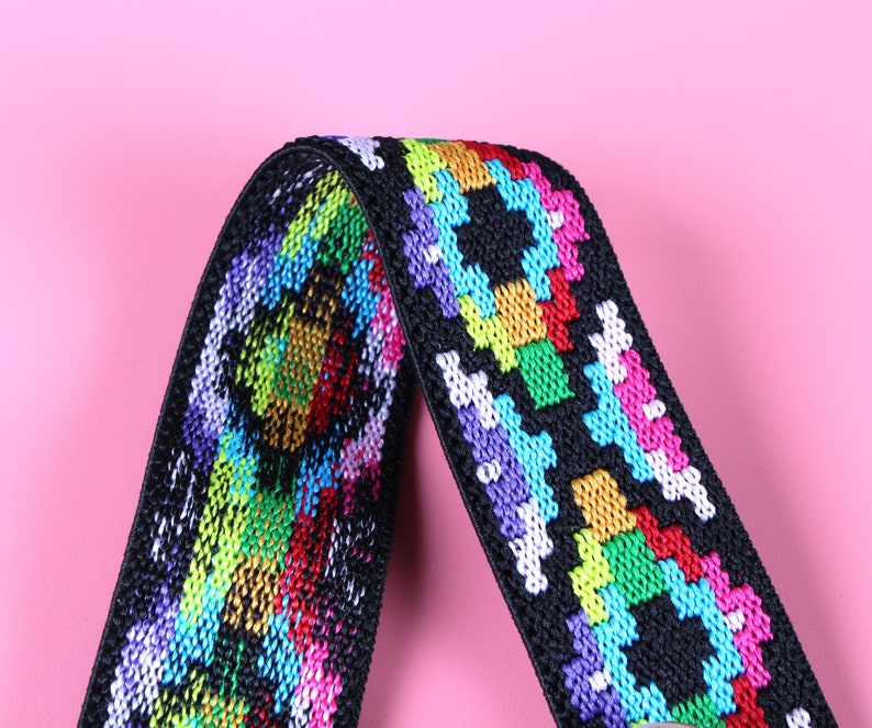 May include: A black elastic strap featuring a woven, multi-colored geometric pattern in vibrant shades of neon pink, bright blue, lime green, yellow, and purple. The fabric has a textured, woven appearance with a repeating, symmetrical design.