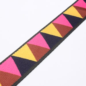 Può includere: Primo piano di un cinturino tessuto con un motivo geometrico ripetuto. Il motivo presenta triangoli nei toni del rosa, giallo, marrone e blu navy, su un bordo nero. Il cinturino viene probabilmente utilizzato per borse o accessori.