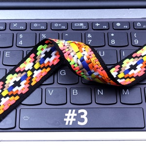 Op de afbeelding: Een kleurrijk geweven band met zwarte, witte, rode, oranje, gele, blauwe en groene accenten. Het band ligt op een zwart laptoptoetsenbord met het nummer "#3" zichtbaar op een van de toetsen.